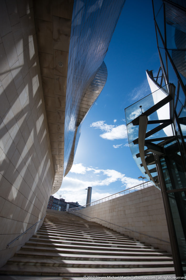 Wide exterior stairs rise beneath a curved titanium overhang between museum walls and glass structure toward blue sky.