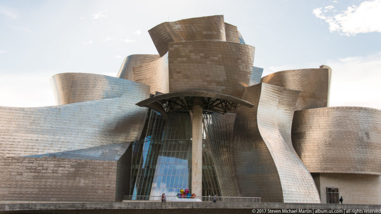 Low frontal view of the museum’s central glass opening beneath a canopy, surrounded by overlapping curved titanium-clad forms.