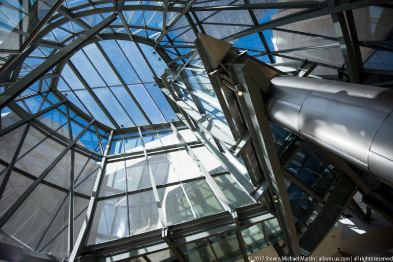Upward view through glass and steel framing in the Guggenheim Museum Bilbao atrium beside a large metallic cylindrical form.