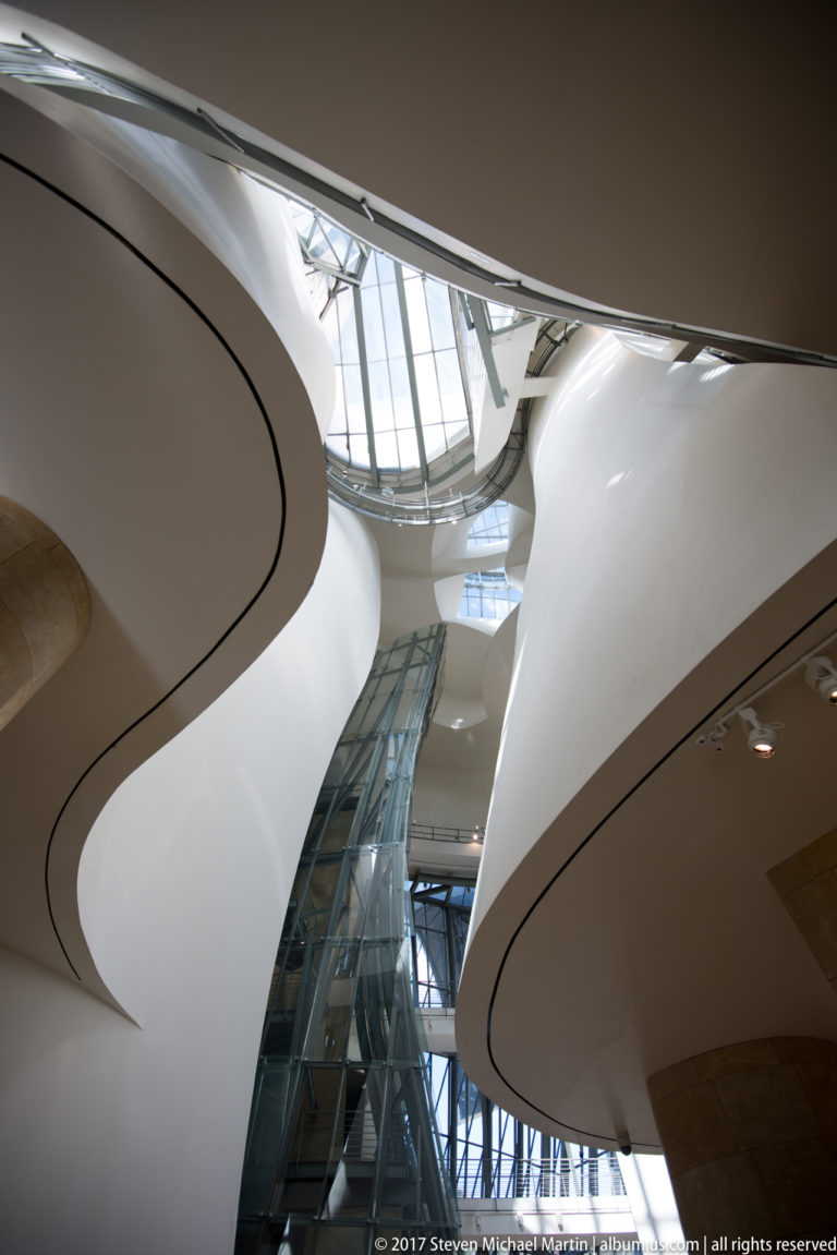 Curving white gallery balconies surround a glass circulation shaft beneath the glazed roof of the Guggenheim Museum Bilbao atrium.