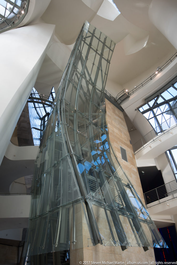Tall glass-and-steel circulation structure rising among white galleries and pale stone walls inside the Guggenheim Museum Bilbao atrium.