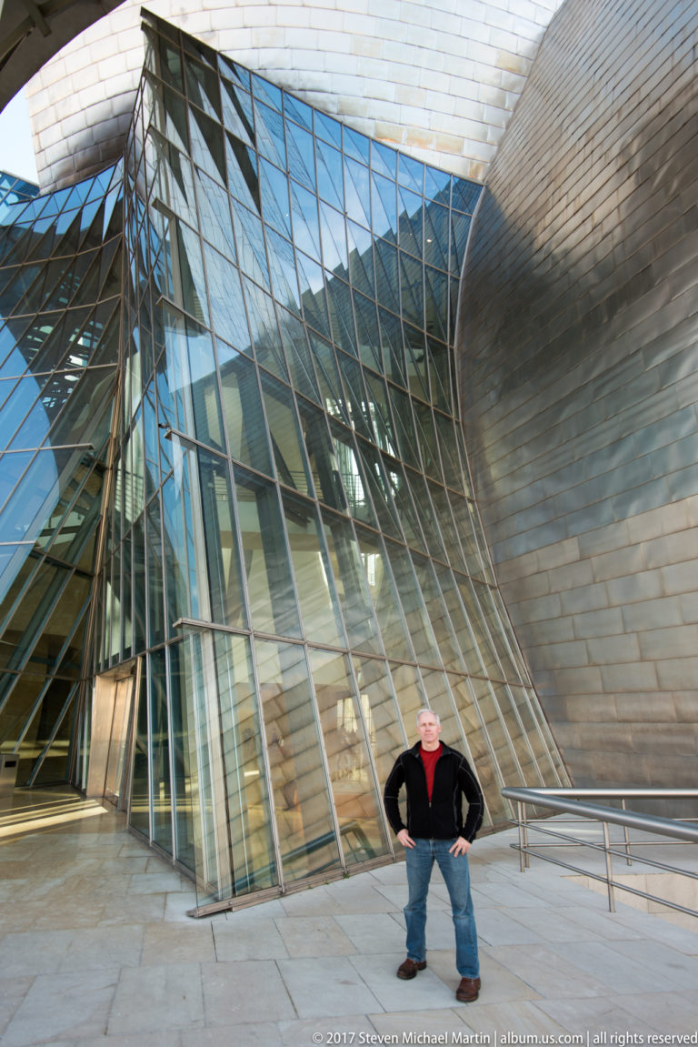 Visitor standing before a tall angled glass façade and curving titanium-clad wall at the Guggenheim Museum Bilbao.