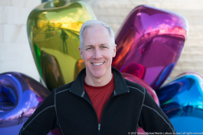 Smiling man in a black jacket posed before the colorful mirrored forms of Jeff Koons’s Tulips in Bilbao.