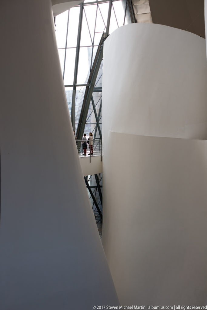 Curving white interior walls form a narrow opening toward glass structure and visitors inside the Guggenheim Museum Bilbao.