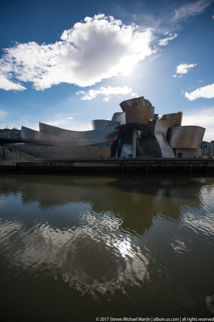 Wide view of the Guggenheim Museum Bilbao across dark river water beneath bright clouds and blue sky.