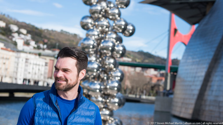 Man in a blue jacket near Anish Kapoor’s reflective sphere sculpture, with the river, bridge, and Guggenheim Museum Bilbao behind.
