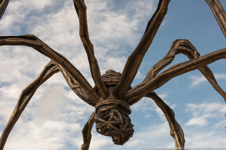 Low-angle close view beneath Louise Bourgeois’s giant spider Maman, with its body and long jointed legs against the sky.