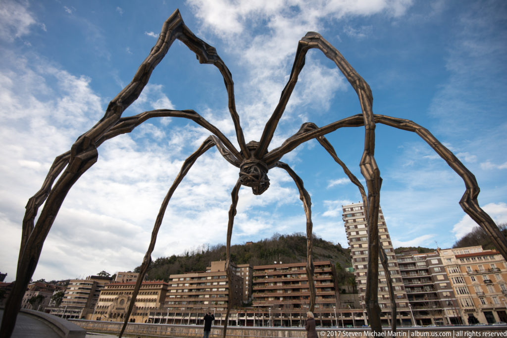 Wide low-angle view of Louise Bourgeois’s giant spider Maman with Bilbao apartment buildings and hillside beyond.
