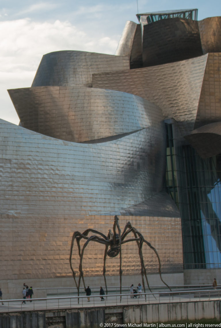 Giant spider sculpture silhouetted against sweeping titanium museum walls and a tall glass bay, with visitors on the promenade.