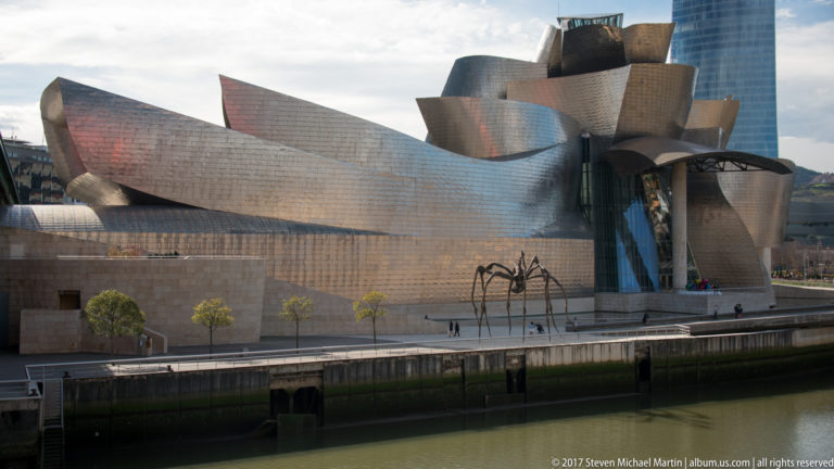 Elevated view of the titanium-clad Guggenheim Museum Bilbao with the giant spider Maman on the riverside promenade below.