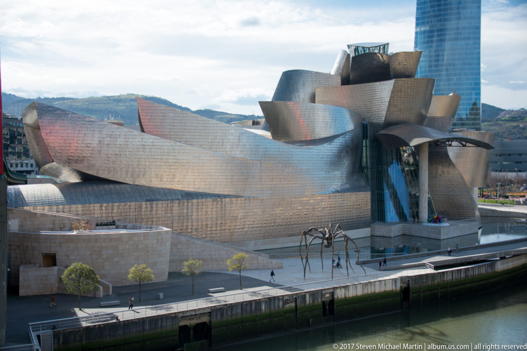 Elevated oblique view of the Guggenheim Museum Bilbao, with Maman on the promenade and a tall glass tower behind.