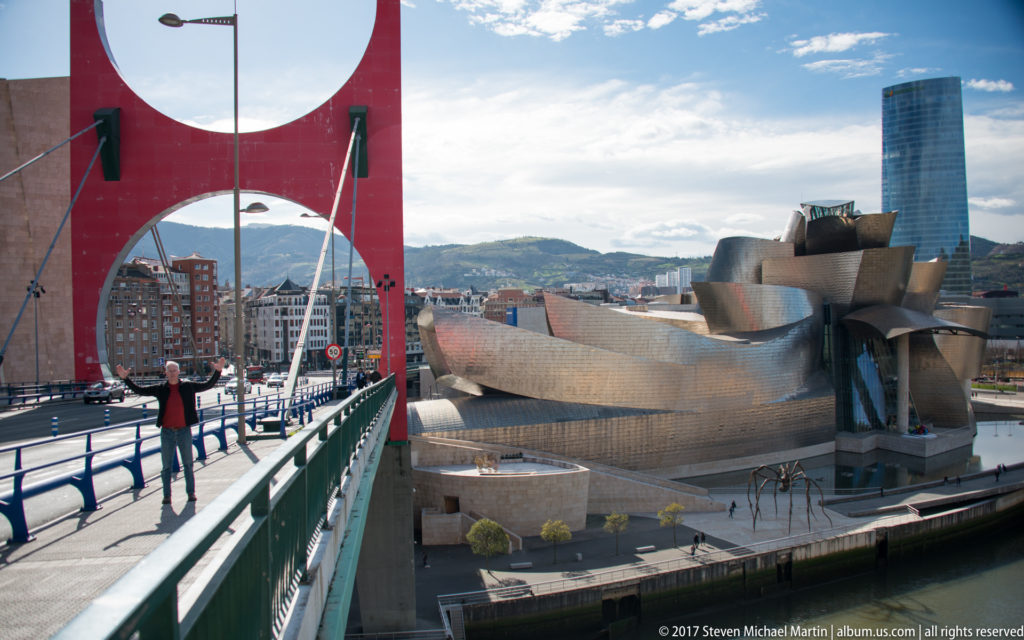 Red bridge portal and pedestrian walkway frame the Guggenheim Museum Bilbao below, with a tall glass tower and hills beyond.