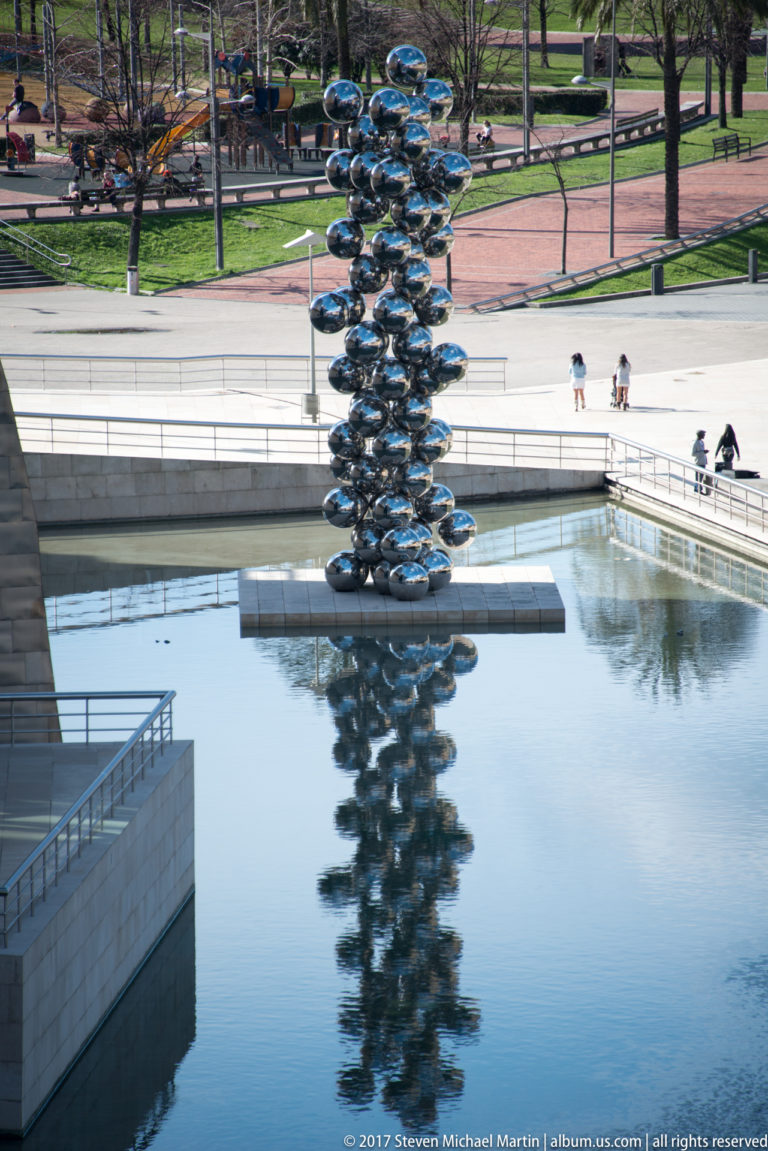 Reflective stacked-sphere sculpture stands in a shallow pool, mirrored in the water beside walkways and a landscaped park.