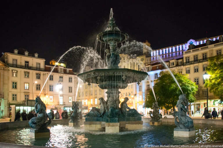 Illuminated tiered fountain with sculpted figures and arcing water jets in Rossio Square at night.