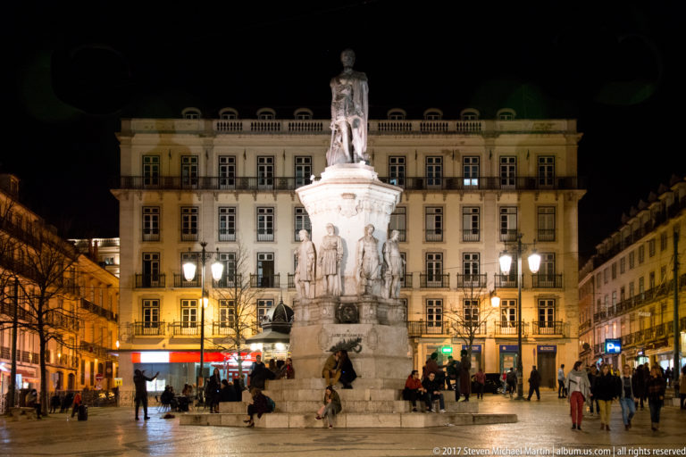 Tall monument to Luís de Camões with standing poet above smaller figures, illuminated in a busy Lisbon square at night.
