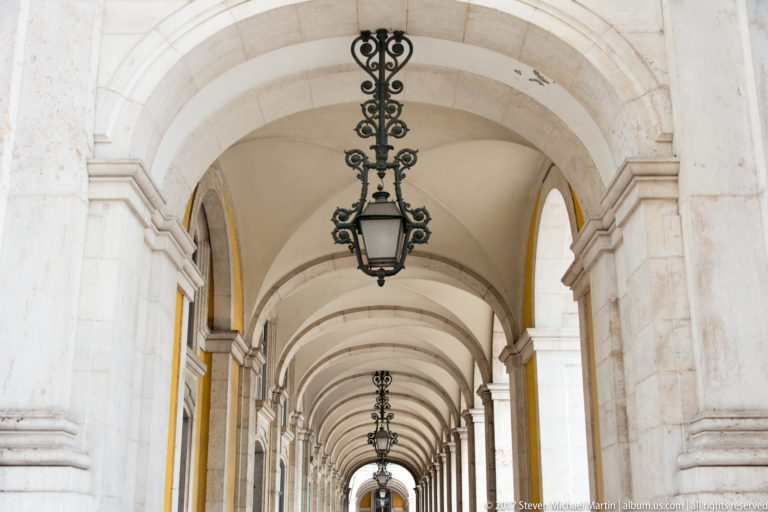 Long arcaded gallery at Praça do Comércio with repeated stone vaults and ornate black hanging lanterns.