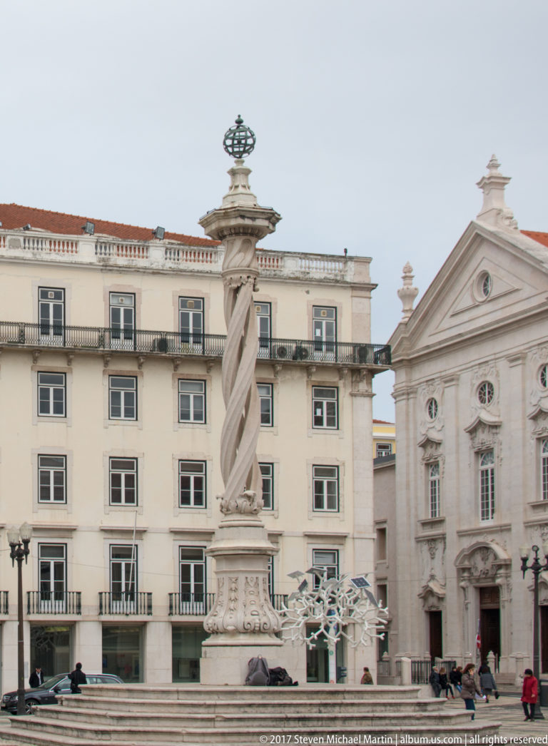 Tall twisted stone column with metal finial in a Lisbon square, with pale buildings and a church façade behind.
