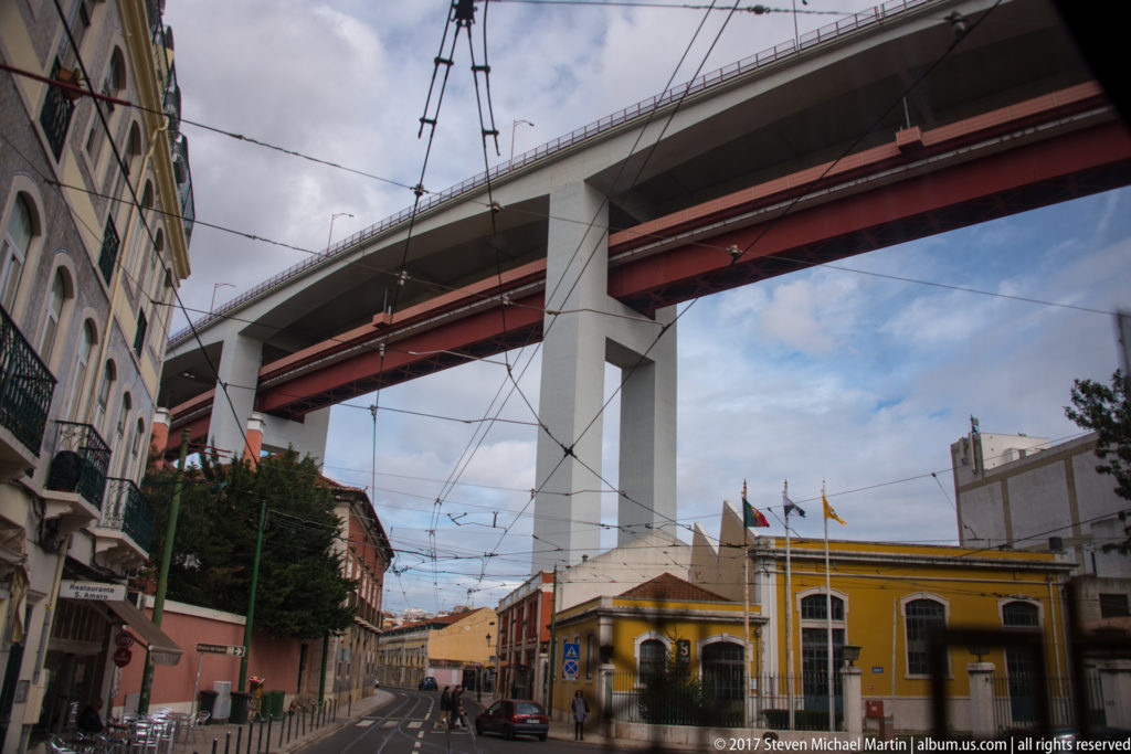 Elevated concrete roadway with red underside spanning a narrow Lisbon street lined by older buildings and overhead wires.