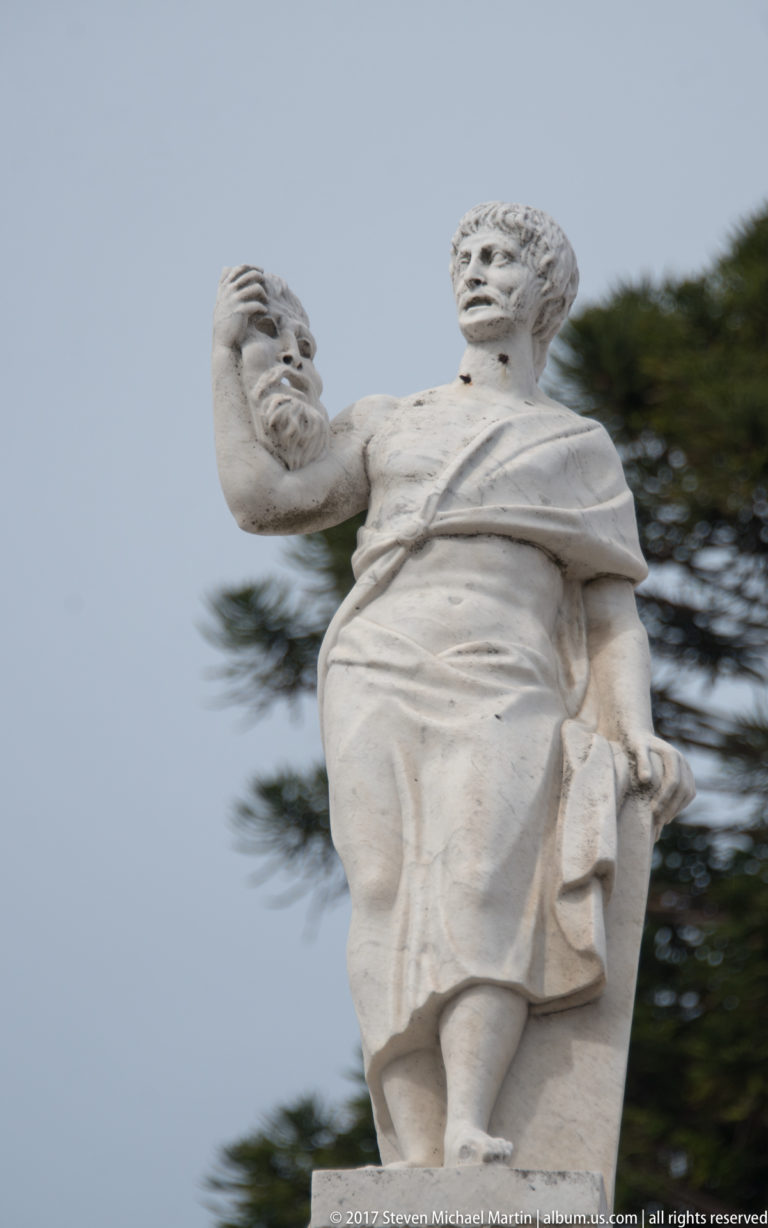 Pale stone statue of a draped male figure raising a rounded object against sky and trees.