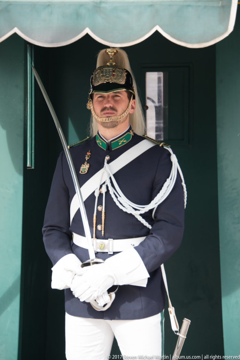 Ceremonial guard in dark uniform, white gloves and plumed helmet holding a curved sword before a green sentry box.