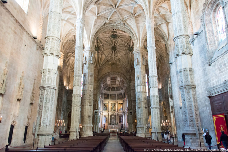 Jerónimos Monastery church nave with slender carved piers, intricate rib vaults, pews, and chancel beyond.