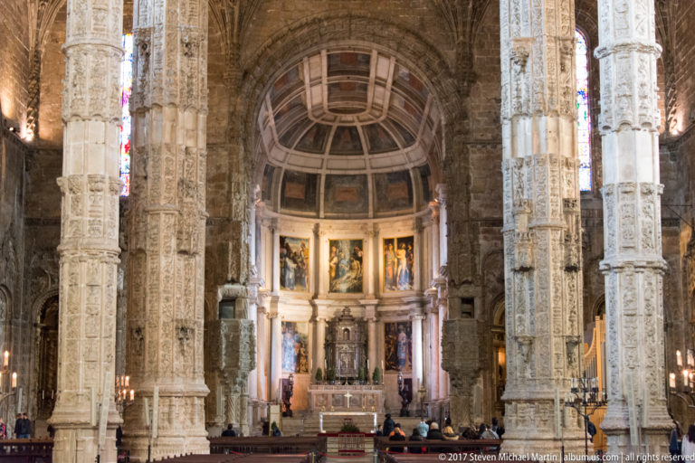 Main chapel of the Jerónimos Monastery church framed by carved piers, with paintings and altar beneath a rounded coffered apse.