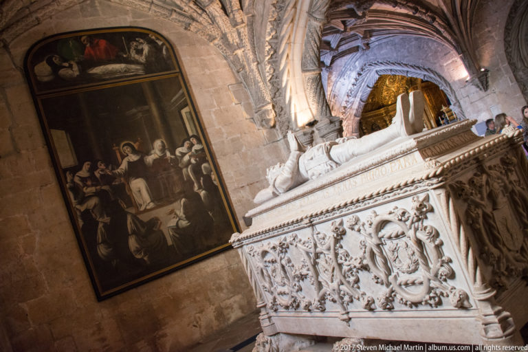 Ornate pale stone royal tomb with recumbent effigy and heraldic carving beside a large religious painting in Jerónimos church.