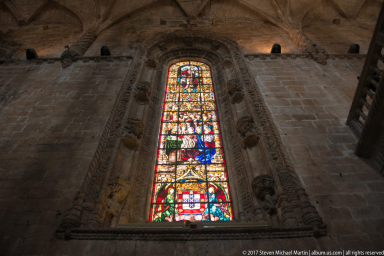 Tall stained-glass window with religious figures and Portuguese heraldry set in the stone wall of the Jerónimos church.