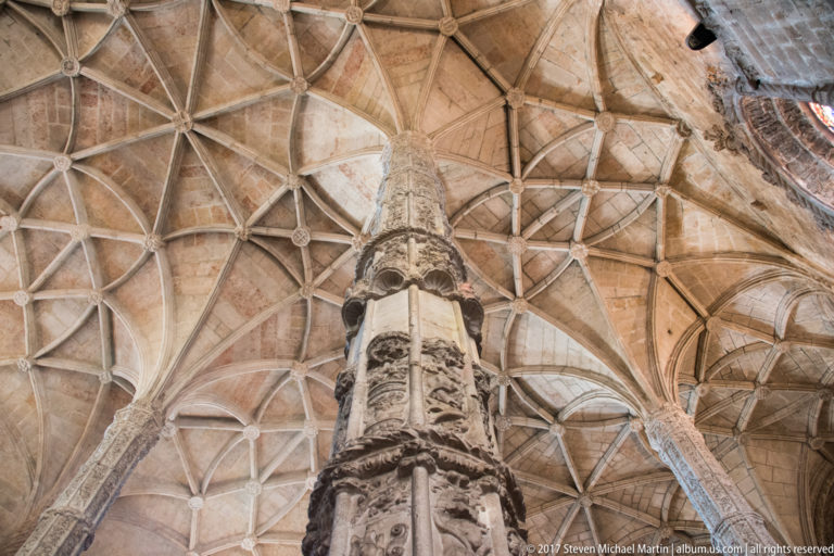 Upward view of intricate stone rib vaulting springing from a richly carved pier inside the Jerónimos Monastery church.