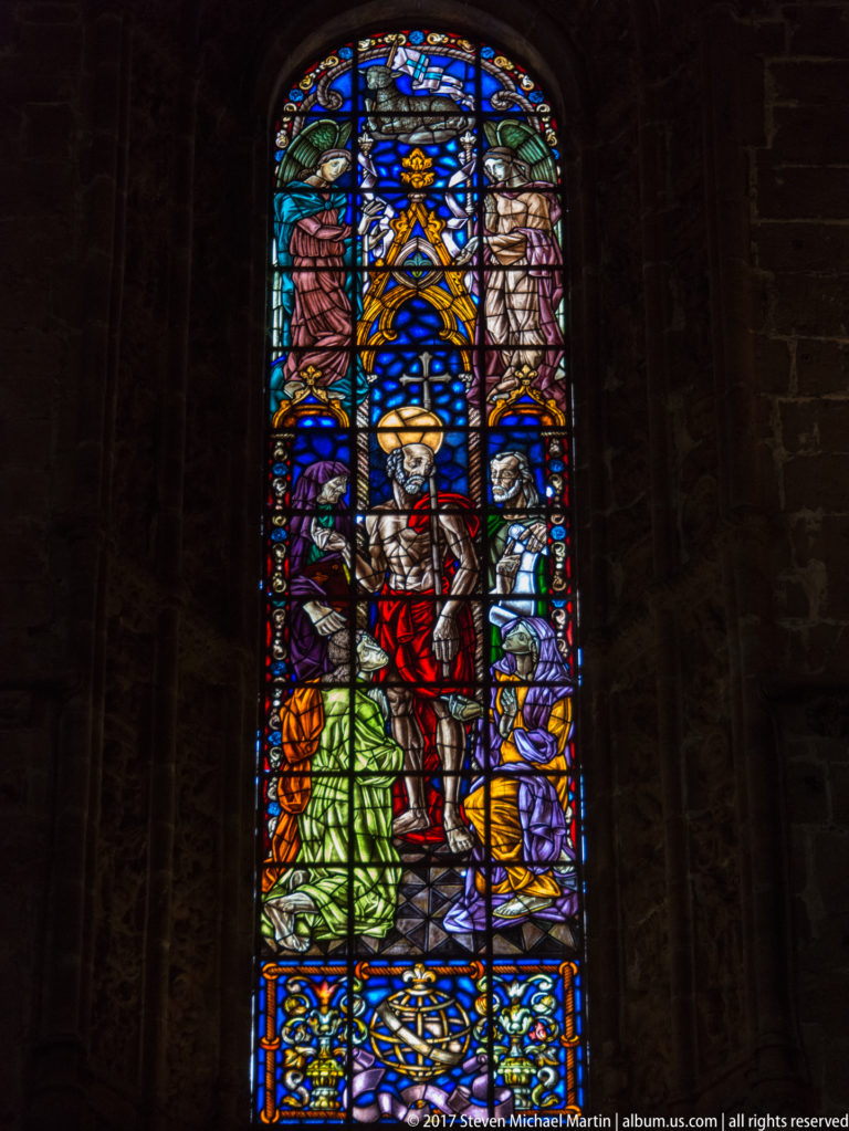 Figurative stained-glass window with central standing figure, kneeling figures, and vivid blue, red, green, and gold glass.