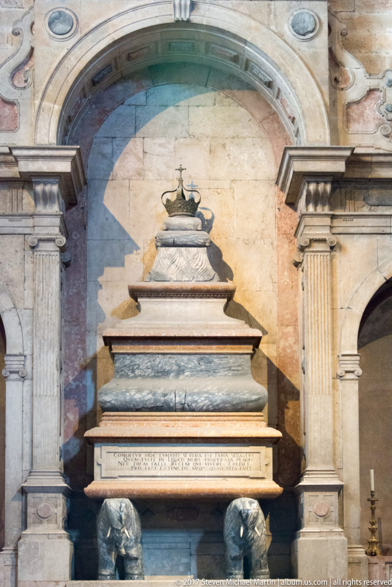 Tiered pale stone tomb monument with inscription and dark sculptural supports in an arched bay inside Jerónimos church.