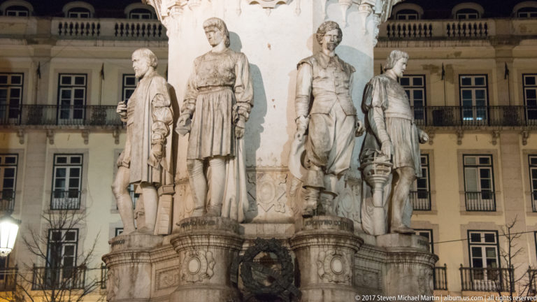 Close view of several carved standing figures arranged around the stone base of the Camões monument.