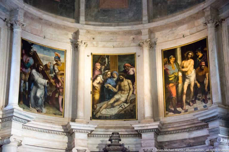 Three large religious paintings arranged around a curved pale-stone chapel wall inside the Jerónimos Monastery.