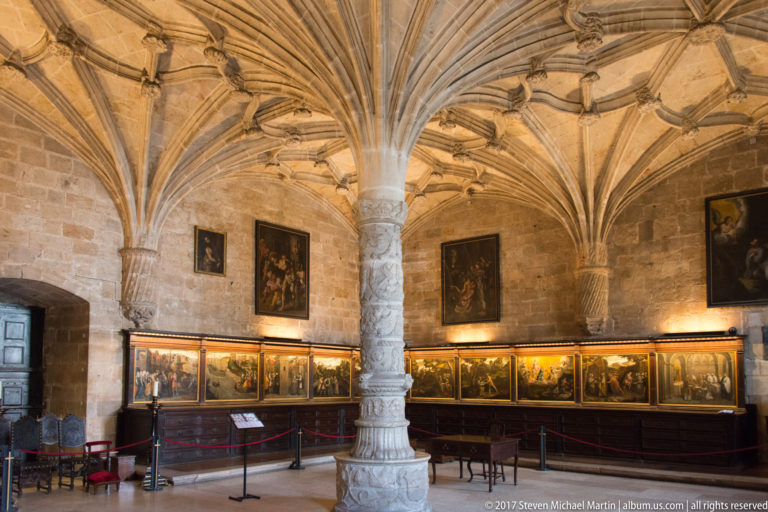 Rib-vaulted stone chamber in the Jerónimos Monastery with large paintings above a continuous row of smaller narrative panels.