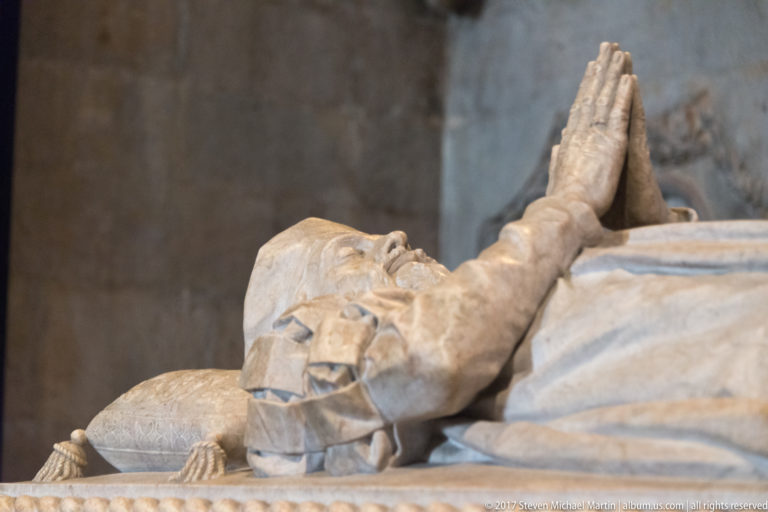 Close view of folded praying hands and draped sleeves on a recumbent stone tomb effigy.