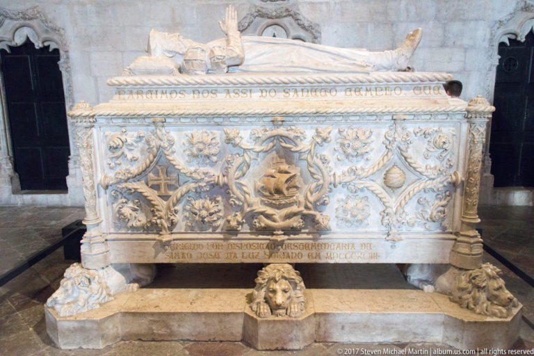 Elaborate pale-stone tomb with recumbent effigy, heraldic carving, inscriptions, and lion-head supports in the Jerónimos Monastery.