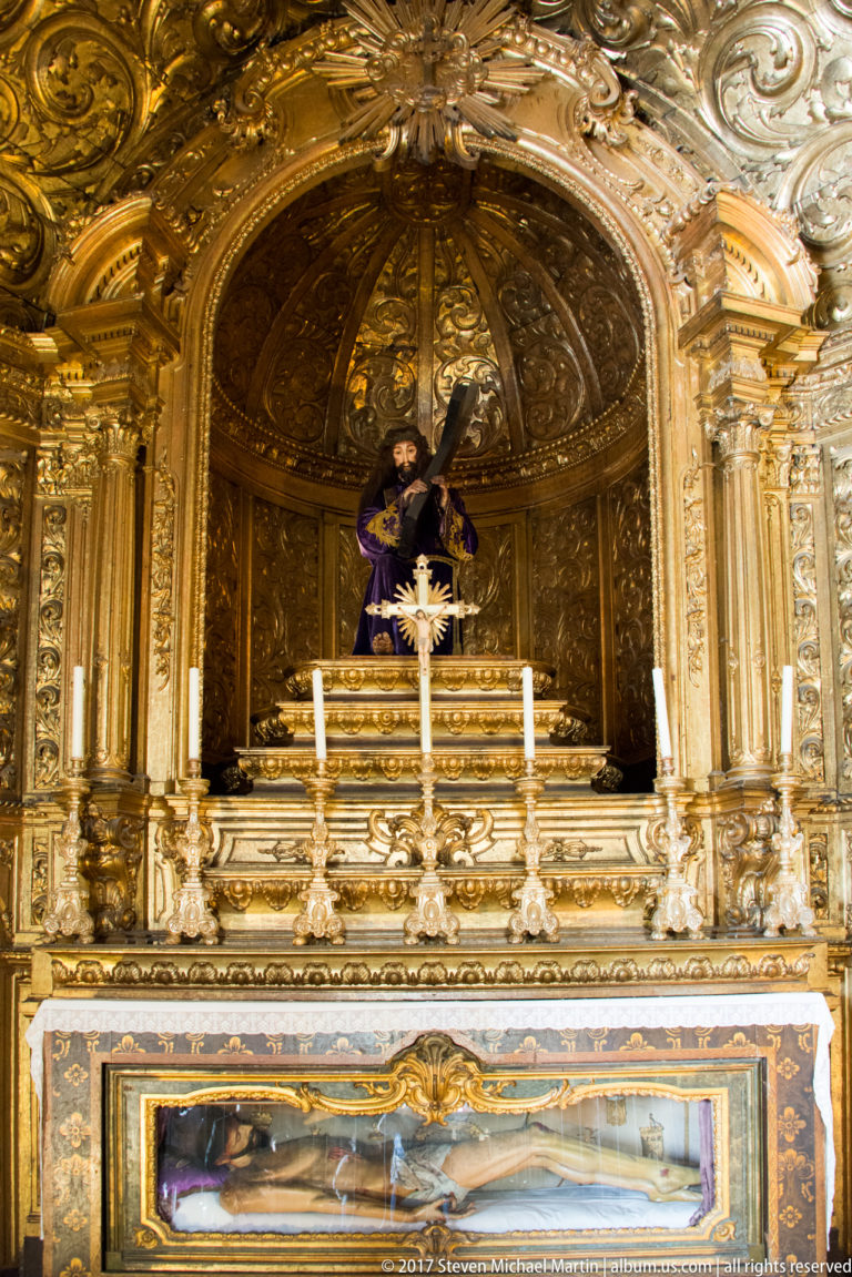 Gilded Baroque chapel altar with dark-robed devotional figure above and a recumbent figure displayed in a glass-fronted case below.