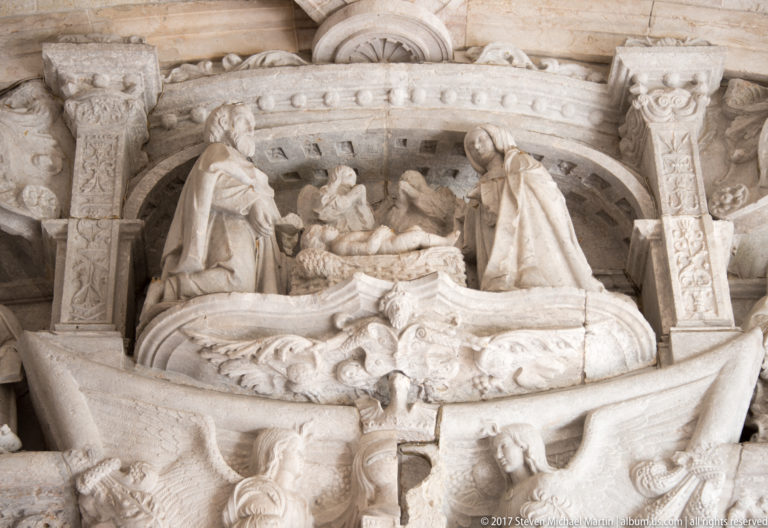 Pale stone Nativity relief with the infant Jesus between Mary and Joseph beneath a carved architectural canopy.