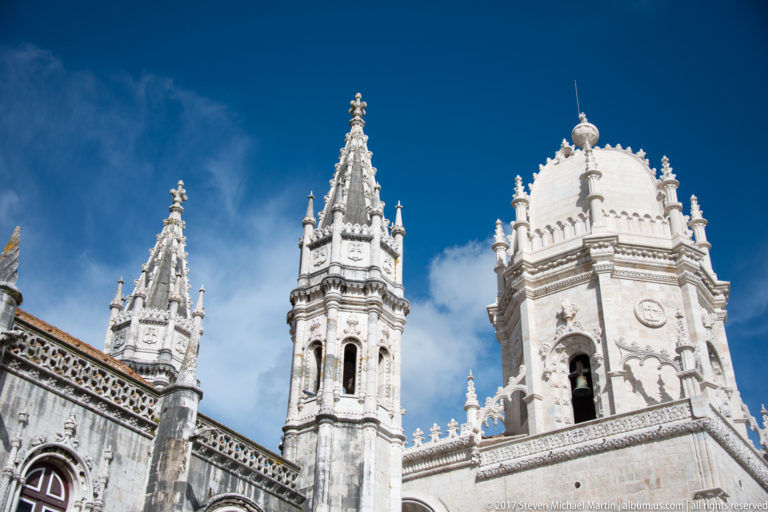 Upward view of the pale limestone towers and pinnacles of the Jerónimos Monastery against a deep blue sky.