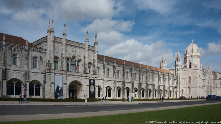 Long pale limestone façade of the Jerónimos Monastery complex with arcades, towers, pinnacles, and a red-tiled roof.