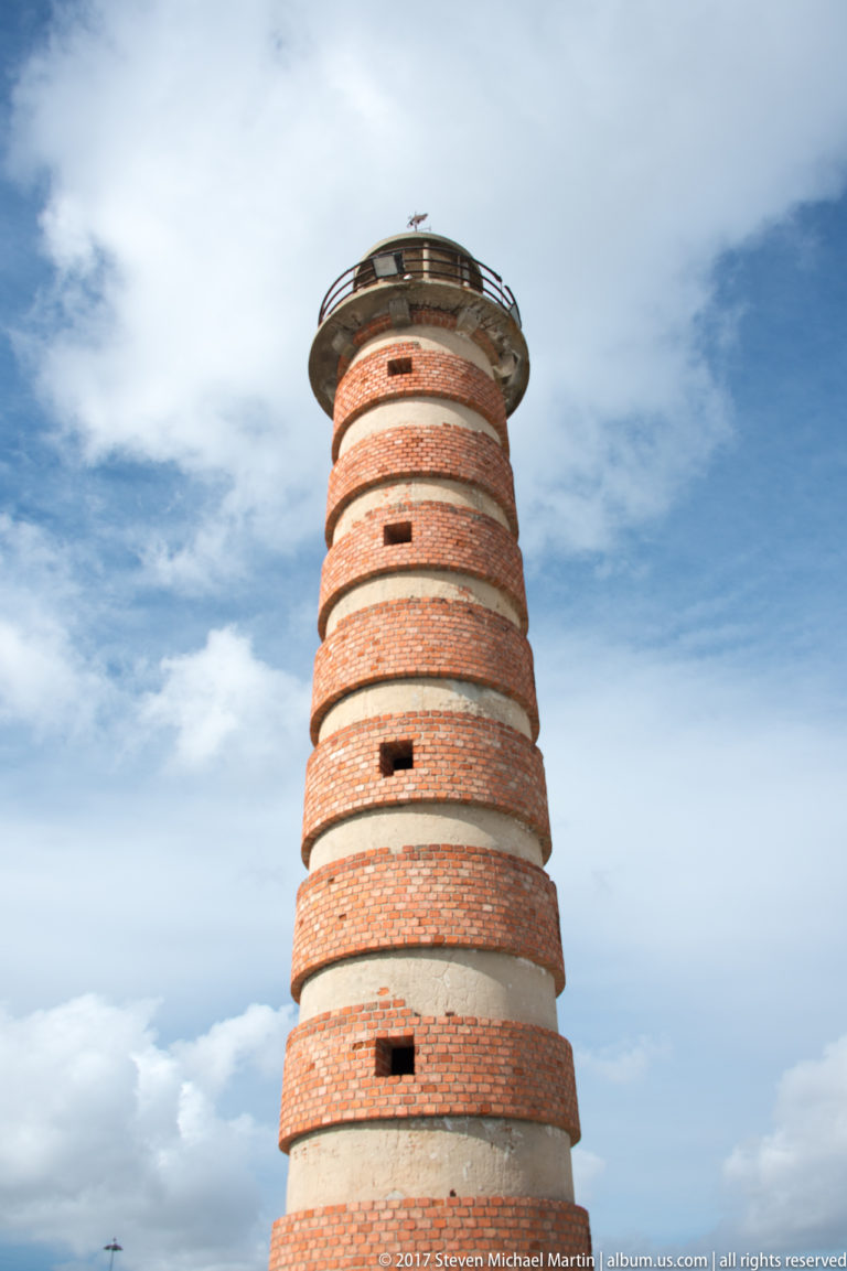 Tapered red-brick cylindrical tower with pale horizontal bands, small openings, and a metal-railed platform at the top.