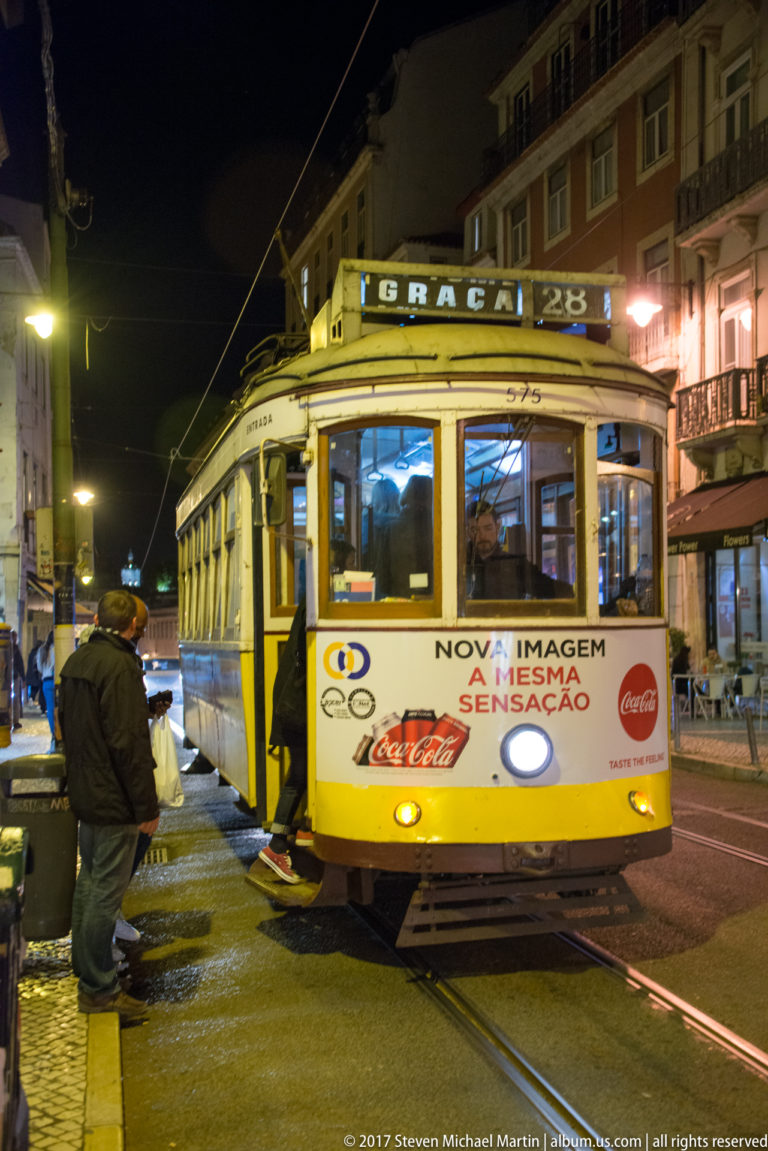 Yellow-and-white Lisbon route 28 tram stopped on tracks at night beneath overhead electric wires.