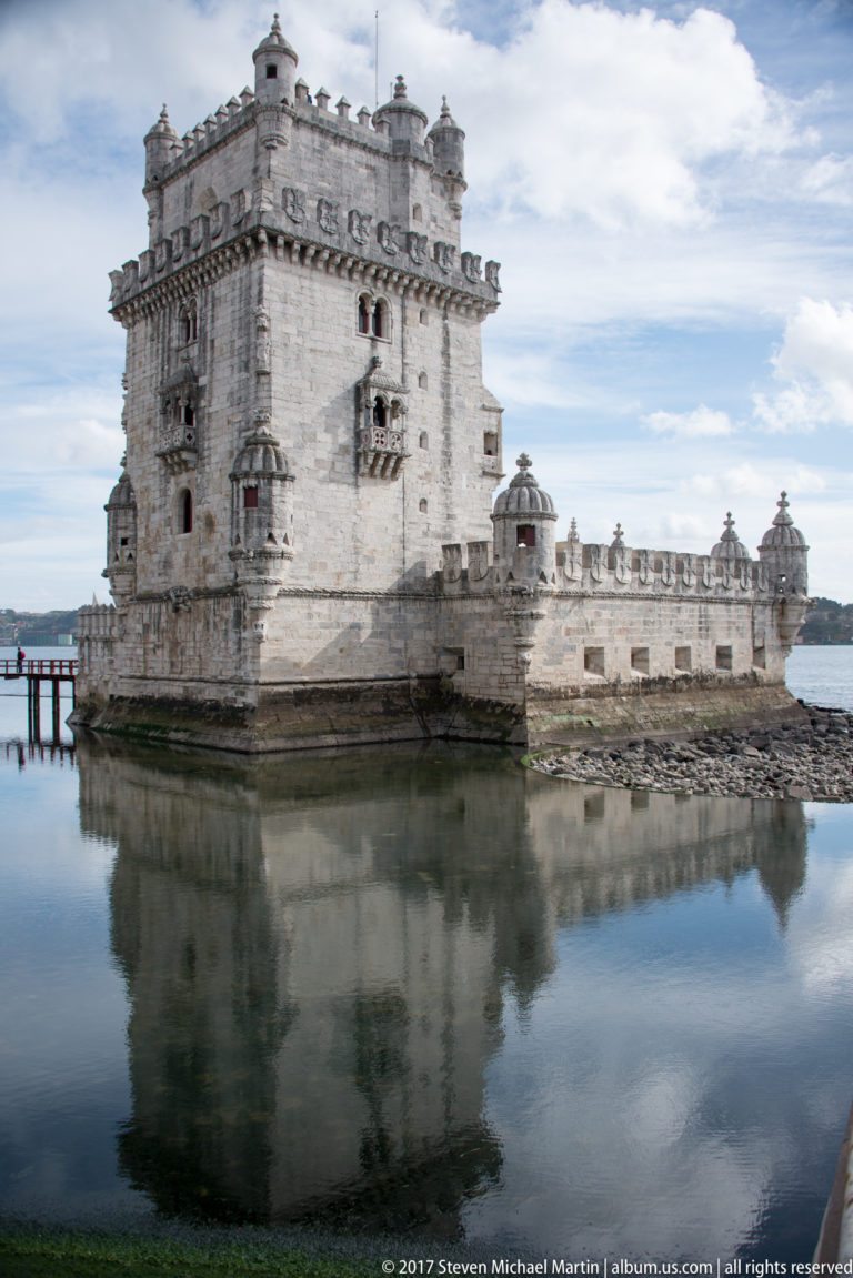 Belém Tower reflected in calm Tagus water, showing the square keep, lower bastion, turrets, and carved stonework.