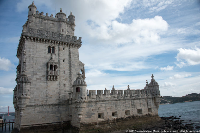 Side view of Belém Tower with square keep, crenellated riverside bastion, corner turrets, and the Tagus beyond.