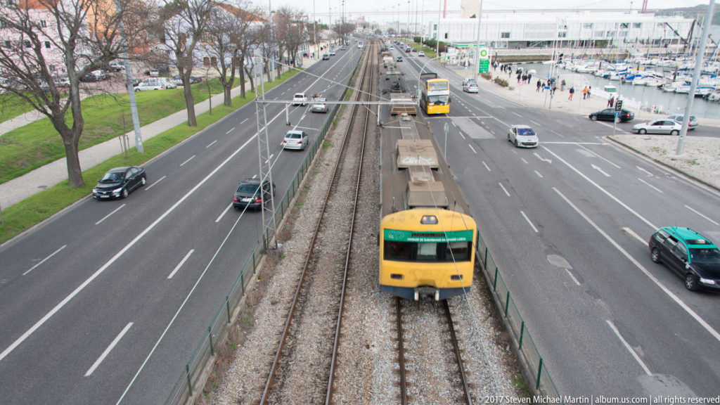 Yellow-and-green commuter train approaching on tracks between busy roads, with marina masts and pedestrians beside the Belém waterfront.