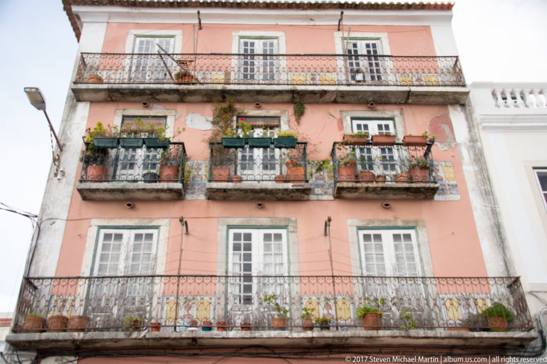 Pink residential façade with wrought-iron balconies filled with potted plants, flowers, and white-trimmed windows.