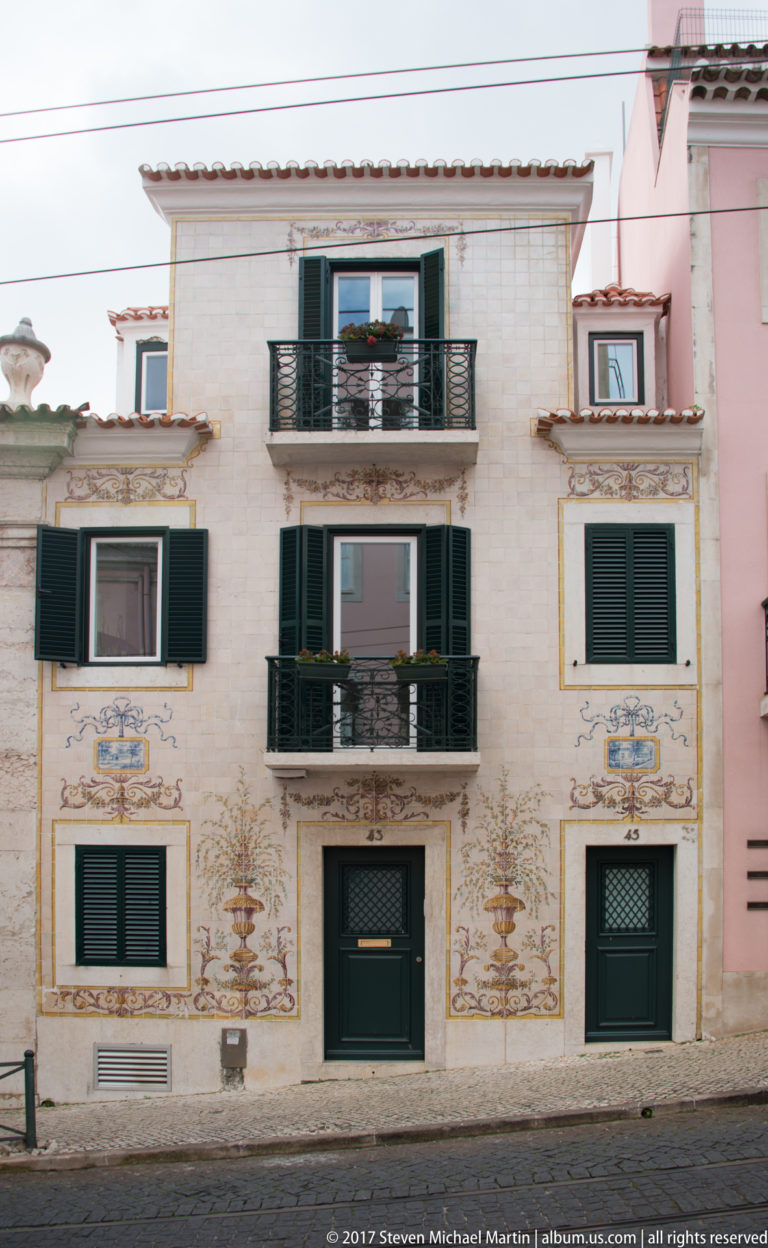 Pale Lisbon residence with dark green shutters, black iron balconies, and decorative floral and scroll motifs on the façade.