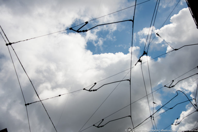 Overhead electric transport wires and junction fittings silhouetted against blue sky and large white clouds.