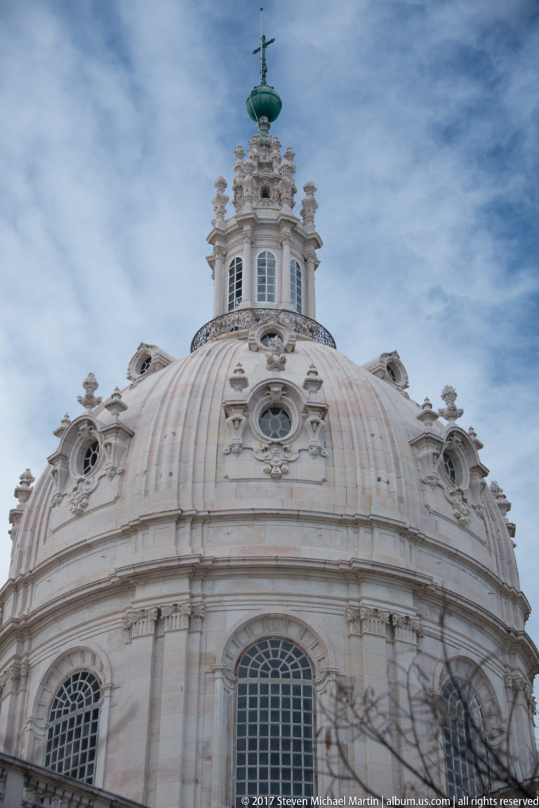 Pale stone dome of the Basílica da Estrela with oval windows, sculpted finials, lantern, and cross against the sky.