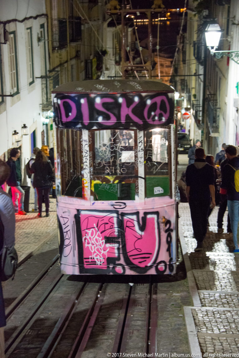 Graffiti-covered funicular car on steep street rails with pedestrians walking alongside in central Lisbon.